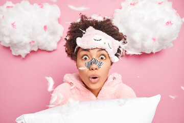 Morning time skin care routine concept. Surprised curly haired young woman wears sleepmask patch on nose dressed in sleep costume holds pillow flying feathers around poses against white clouds