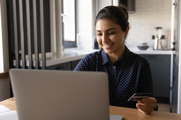 Close up smiling Indian woman paying online, using laptop, holding plastics credit card, sitting at table, happy satisfied customer shopping, making secure internet payment, browsing banking service