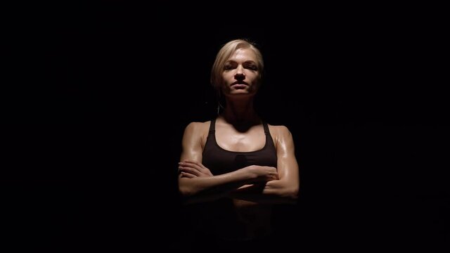 Athletic woman stands in darkness and puts hands on chest, looking at the camera. Sports woman puts arms on chest in a black background studio.