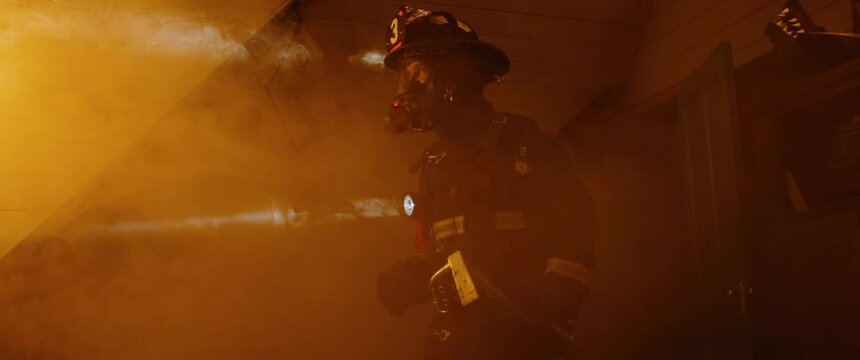 HANDHELD Dramatic shot of American firefighter in full gear walking through smoke and fire in a burning house room. Shot with 2x anamorphic lens. 100 FPS slow motion