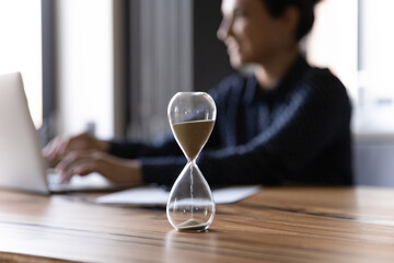 Close up hourglass measuring time, standing on wooden office table, Indian businesswoman working on...