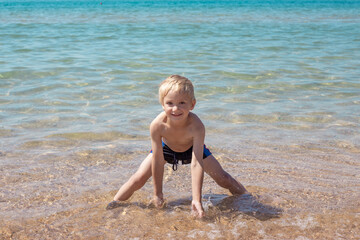 Little blond boy in swimming trunks is playing on the seashore with sand and water, smiling, standing in the water. Summer happy vacation.