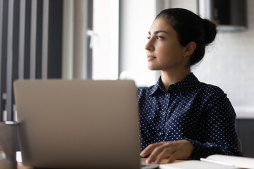 Close up pensive Indian businesswoman looking to aside, pondering strategy, sitting at table with...