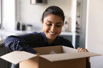 Close up smiling Indian young woman opening cardboard box with awaited online store goods at home, overjoyed satisfied customer addressee received parcel, quick postal delivery service concept - obrazy, fototapety, plakaty