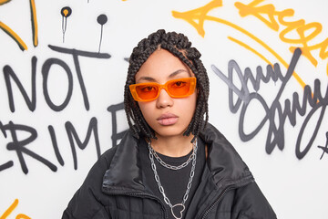 Photo of serious teenage hipster girl with dreadlocks wears trendy orange sunglasses and black jacket looks directly at camera has calm expression belongs to hooligan company of friends. Swag - obrazy, fototapety, plakaty