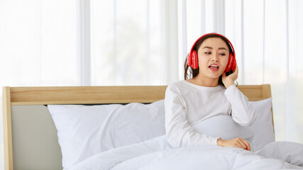 Portrait shot of a young adult Asian woman in casual white sings a song, lying down on the bed and listening to the music with a red headphone. There is copy space at the side of picture