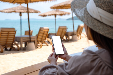 Fototapeta premium Mockup image of a woman holding mobile phone with blank desktop screen while sitting on the beach