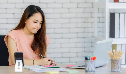 portrait of young Asian businesswoman wearing casual dress sitting on a desk holding pen busy working and writing notes during communicating with laptop in the office