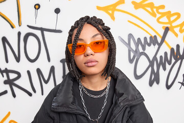 Serious trendy hipster girl wears orange sunglasses black jacket and metal chains around neck belongs to youth subculture poses against graffiti wall. Millennial people style hooliganism concept