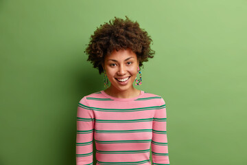 Portrait of lovely young African American looks with friendly happy expression at camera expresses positive emotions being in good mood dressed casually isolated over green vivid background.