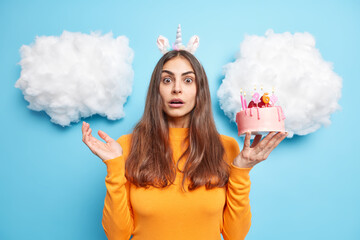 Photo of stupefied brunette woman gazes surprisingly at camera celebrates 26th birthday olds festive cake wears orange jumper isolated over blue background with clouds above. Oh no how many congrats