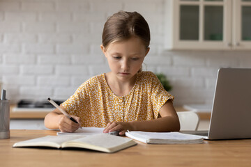 Smart little Caucasian schoolgirl sit at table at home write in notebook do homework assignment learn distant with computer. Small girl child handwrite study online on laptop. Education concept.