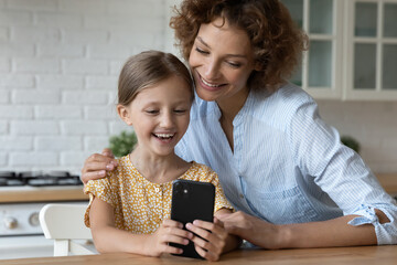 Smiling young Caucasian mother and little 8s daughter look at cellphone screen laugh watch funny...