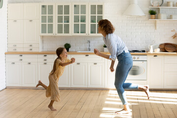 Positive happy young mom and little 8s daughter have fun dance in renovated design kitchen. Smiling overjoyed Caucasian mother and small girl child celebrate relocation to new home. Rental concept.