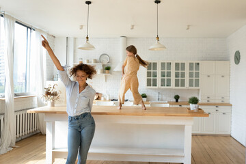 Overjoyed young Caucasian mother and little 8s daughter have fun dancing together in new renovated...