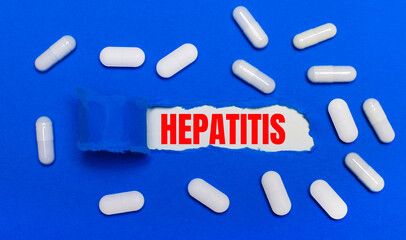 White pills lie on a beautiful blue background. In the center is white paper with the inscription HEPATITIS. Medical concept. View from above.