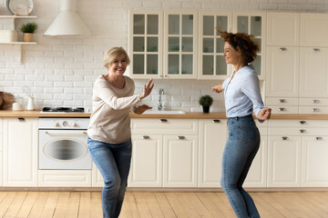 Smiling senior 60s mom and adult millennial daughter dancing together in modern kitchen. Overjoyed mature mother and grownup girl child have fun enjoy family leisure weekend or celebrate relocation.