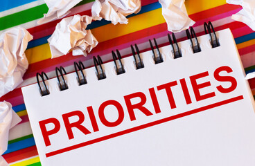 On a multi-colored striped background there are white pieces of paper and a white notebook with the inscription in red PRIORITIES