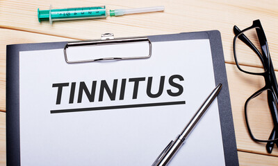 The word TINNITUS is written on a white piece of paper next to black-rimmed glasses, a pen and a syringe. Medical concept