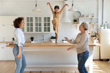 Happy three generations of women dance in new home kitchen have fun together on weekend. Excited smiling little girl with Caucasian mom and grandmother celebrate relocation to own house.