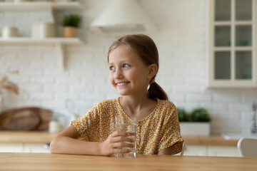 Smiling small Caucasian 9s girl sit at desk at home drink clean crystal mineral still water from glass. Happy little kid child enjoy clear aqua feel dehydrated thirsty. Hydration, diet concept.