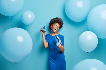 Positive African American woman with curly hair keeps shoes near mouth pretends singing wears dress...