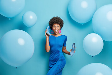 Fototapeta premium Horizontal shot of curly haired young woman wears dress holds high heeled shoes dresses for party poses against blue background with big inflated balloons. People festive event fashion concept