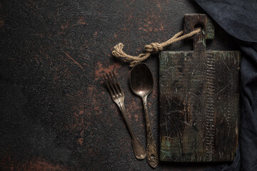 Fototapeta na wymiar Vintage wooden cutting board, cutlery and kitchen towel on dark concrete background. top view with copy space