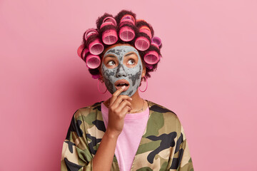 Portrait of surprised housewife applies clay mask on face looks wondered above makes hairstyle with hair rollers wears domestic robe poses against pink background. Beauty procedures at home.