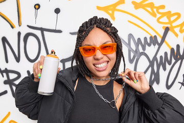 Trendy fashionable teenage girl dressed in black clothes orange sunglasses and metal chain has braid hairstyle poses with aerosol spray makes creative graffiti on street walls. Urban art design - obrazy, fototapety, plakaty