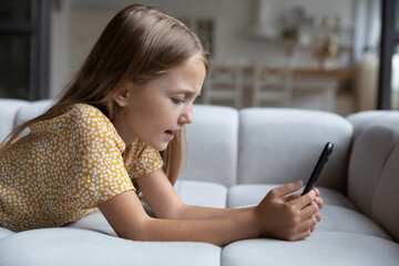 Little Caucasian teen 9s girl child lying on sofa at home look at cellphone screen play game online. Small 8s kid relax in living room use modern smartphone gadget talk speak on video call on device.