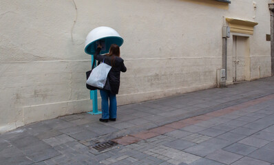 Front view of middle age woman making phone call in phone booth in front of light-colored wall, inside empty street.