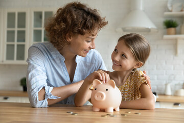 Smiling young Caucasian mom and little daughter put coins money into piggybank mange family budget...