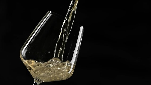 Pouring gold wine into goblet at black background. Close-up of filling wine glass with white wine in super slow motion. White wine forms beautiful wave in glass with copy space
