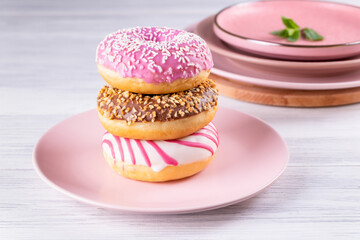 Three iced donuts are stacked on a pink ceramic plate on a light wooden surface with a stack of plates and a cutting board. Selective focus.