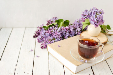 Romantic background with a cup of tea, lilac flowers and a book over a white wooden table. Leisure concept, spring breakfast.