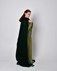 full length portrait of red haired girl wearing celtic, green medieval gown with fantasy velvet cloak. Standing pose isolated against a studio background.