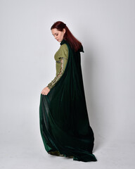 Naklejka premium full length portrait of red haired girl wearing celtic, green medieval gown with fantasy velvet cloak. Standing pose isolated against a studio background.