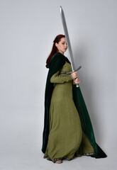 full length portrait of red haired girl wearing celtic, green medieval gown with fantasy velvet cloak. Standing pose isolated against a studio background.