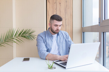 Young man working from the office sitting by the window, at the laptop, dressed in casual clothes businessman solves business on the phone