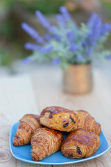 Sweet buns with raisins and croissants on a blue plate and a wooden table and a blurred background, bouquet of lavender in a vase.