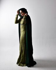 Naklejka premium full length portrait of red haired girl wearing celtic, green medieval gown and velvet cloak with shadowy backlighting. Standing pose isolated against a studio background.