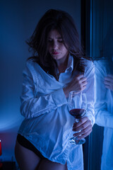 Lonely woman in white man's shirt drinking wine near the window at home