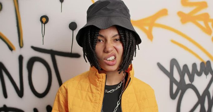 Teenagers lifestyle subculture concept. Fashionable hipster girl with dreadlocks shows golden teeth has behaviour of cool rapper wears hat and jacket poses against graffiti wall. Street urban style