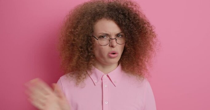 Displeased curly haired young woman points at herself asks who me looks unhappily at camera wears round spectacles and shirt isolated over pink background. Indignant female says no shakes head