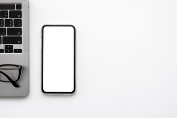 Smartphone with blank mockup screen is on top of white office desk table with laptop computer. Top...