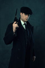 Menacing vintage man in a dark striped suit and a cap holding a handgun in front of a dark wall.