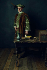 Renaissance man stands behind a table with books on it, a tin pitcher, a tin goblet, a candlestick...