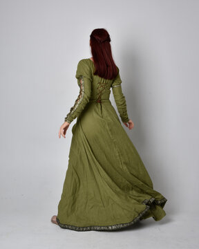 full length portrait of red haired girl wearing celtic, green medieval gown. Standing pose isolated against a studio background.