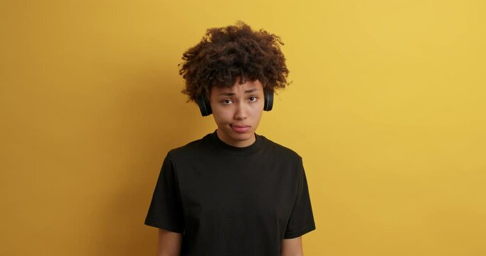 Upset dejected dark skinned woman with curly hair looks sadly at camera being offended by someone listens music to entertain wears wireless headphones black t shirt isolated on yellow studio wall